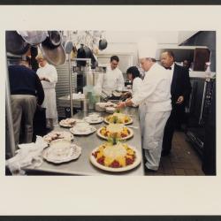 In this photograph, White House Executive Pastry Chef Roland Mesnier plates petits fours for a dinner at Camp David. In the back, White House Executive Chef Walter Scheib and chef Cristeta Comerford stack dishes. 
